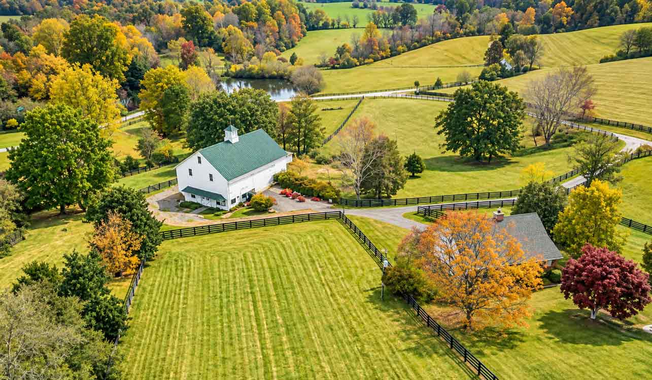 Aerial view of a Nelson County horse farm with a white barn, fenced pastures, rolling fields, mature trees, and a pond in Central Virginia